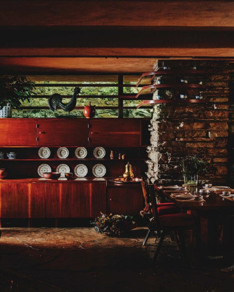 A dimly lit interior view of Frank Lloyd Wright's Fallingwater house, showing a wooden dining sideboard with a display of plates, and a table set with chairs in the foreground.