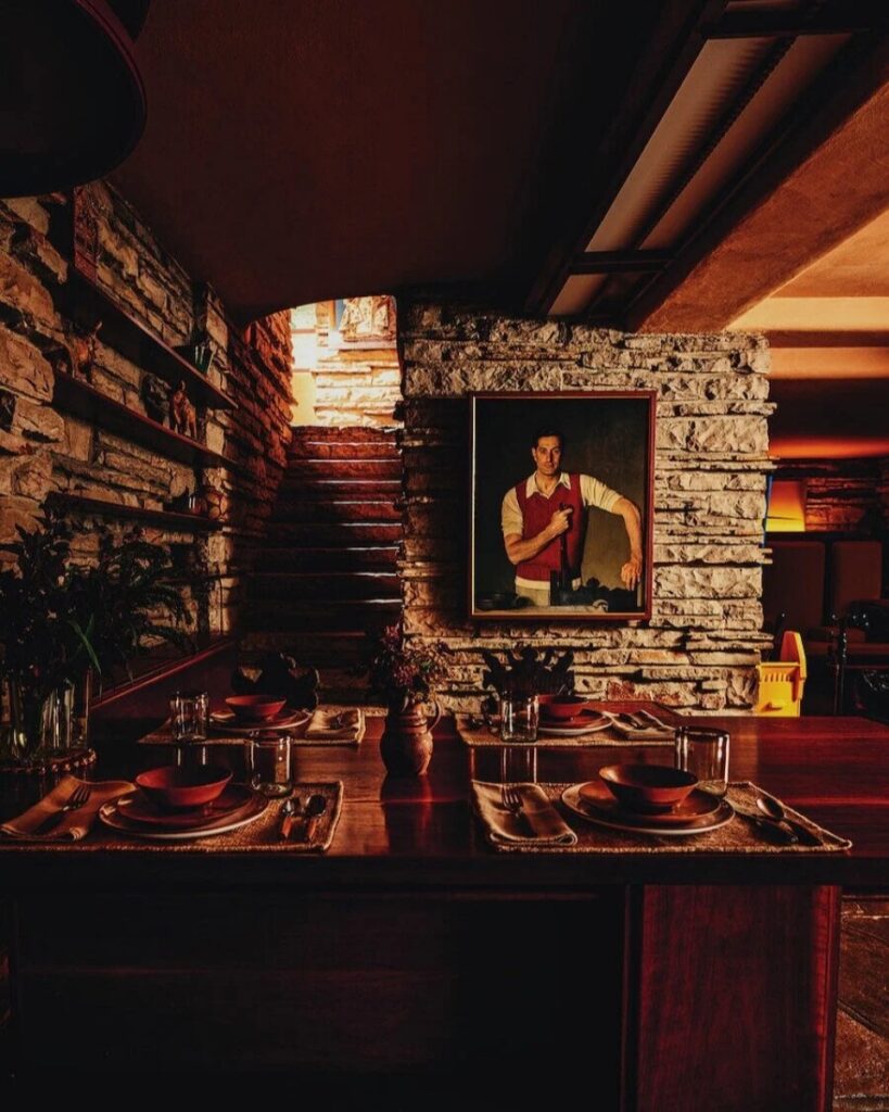 A painting of Edgar J. Kaufmann, Sr. in a red vest, hanging on a stone wall in the dining area of Frank Lloyd Wright's Fallingwater, with a wooden table set for a meal in the foreground.