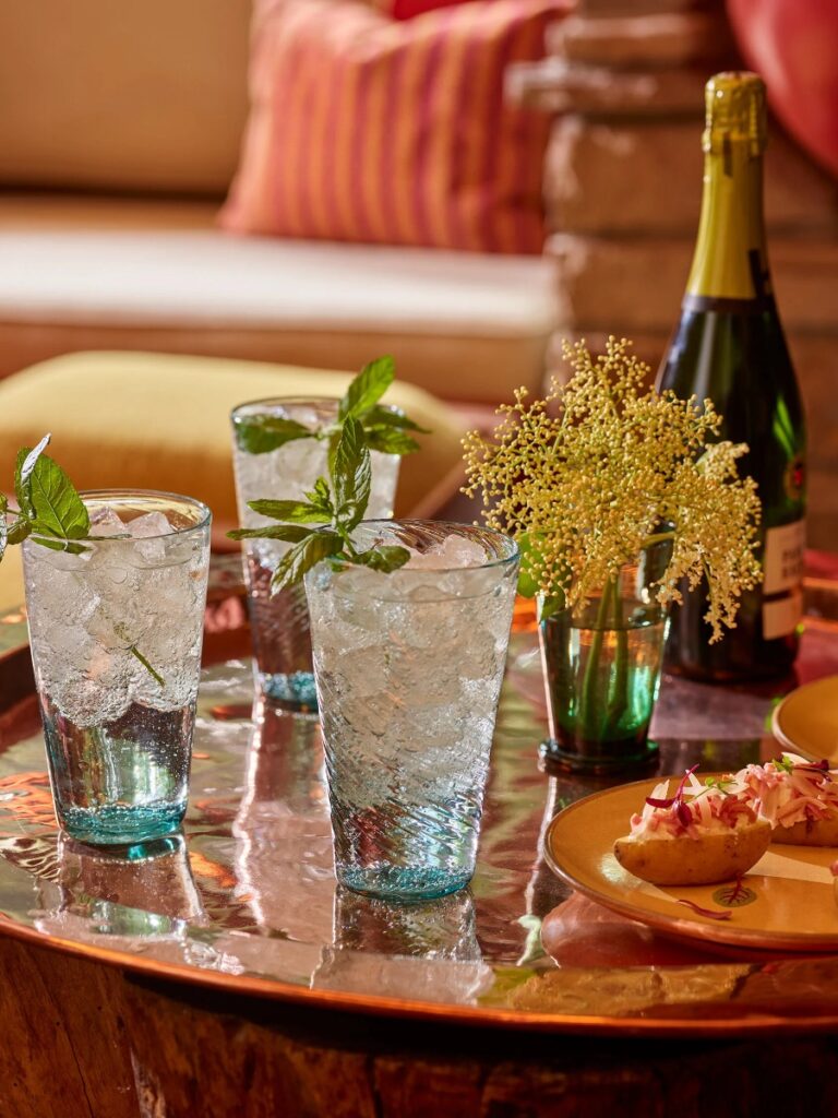 A Hugo cocktail with fresh mint and crushed ice on a tray, with other glasses and a bottle of sparkling wine in the background.