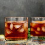 A moody shot of two glasses of Ancho Chile Old Fashioned cocktails on a bar, with scattered sugar crystals on the surface.