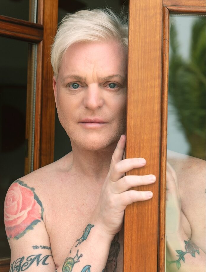 A man stands behind a wood beam shirtless with white hair.