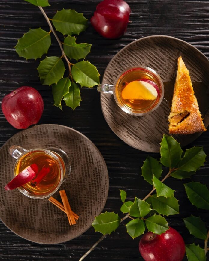 A warm Apple Cinnamon Hot Toddy in a clear mug, garnished with an apple slice and a cinnamon stick, served with a slice of cake on a brown plate.