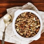 A large white bowl filled with homemade apple cinnamon spice granola, placed on a beige napkin with a silver spoon on a wooden table.