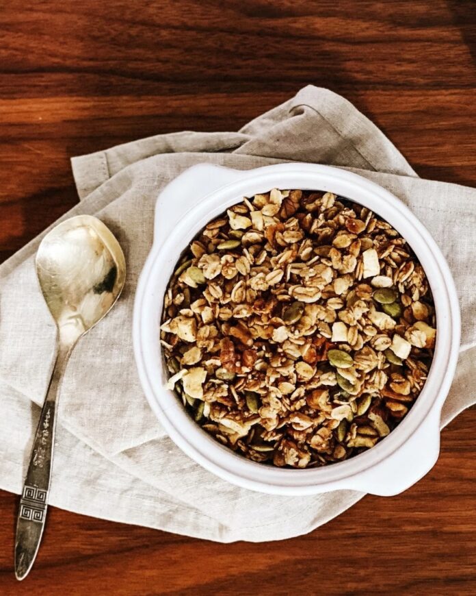 A large white bowl filled with homemade apple cinnamon spice granola, placed on a beige napkin with a silver spoon on a wooden table.
