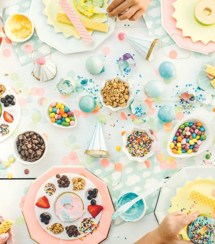 A colorful overhead view of a table with plates of sliced apples decorated to look like donuts with various toppings, with bowls of candy and sprinkles, and children's hands reaching for the food.