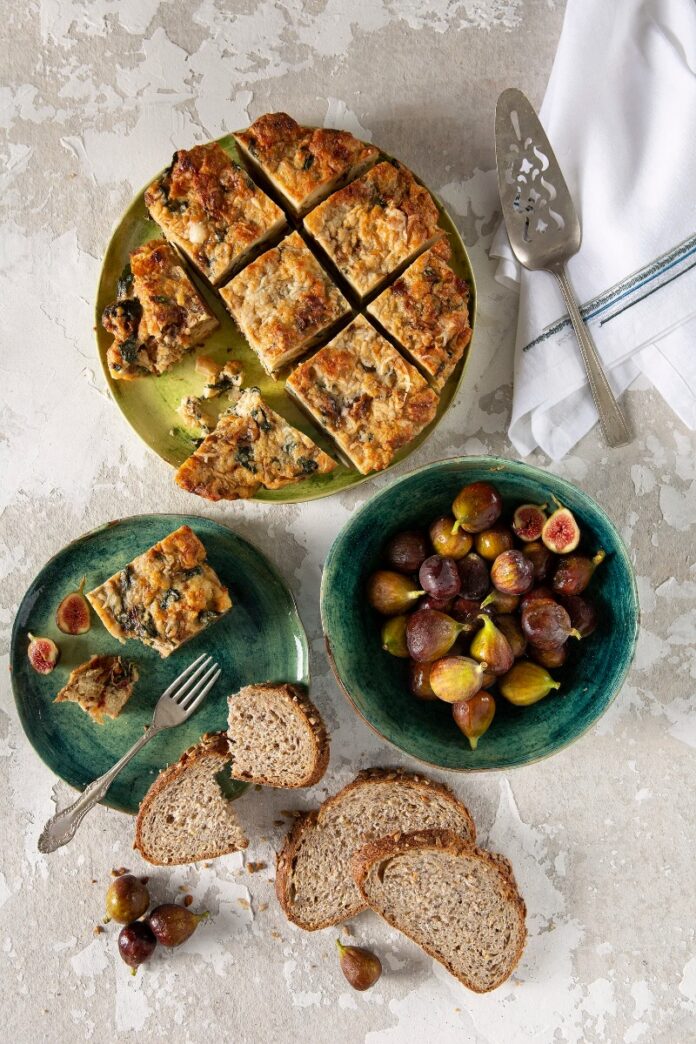 Breakfast Strata with Caramelized Onions, Dates and Gruyere A table set with a delicious breakfast casserole, complemented by bread, figs, and an assortment of fresh fruits for sharing.
