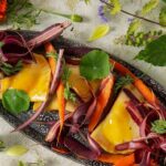 A plate of carrot pierogies garnished with carrot ribbons and fresh herbs, surrounded by flowers and green and red vegetables.