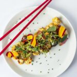 A plate of cauliflower stir-fry with sliced ginger, carrots, and green onion, with a pair of red chopsticks resting on the side.
