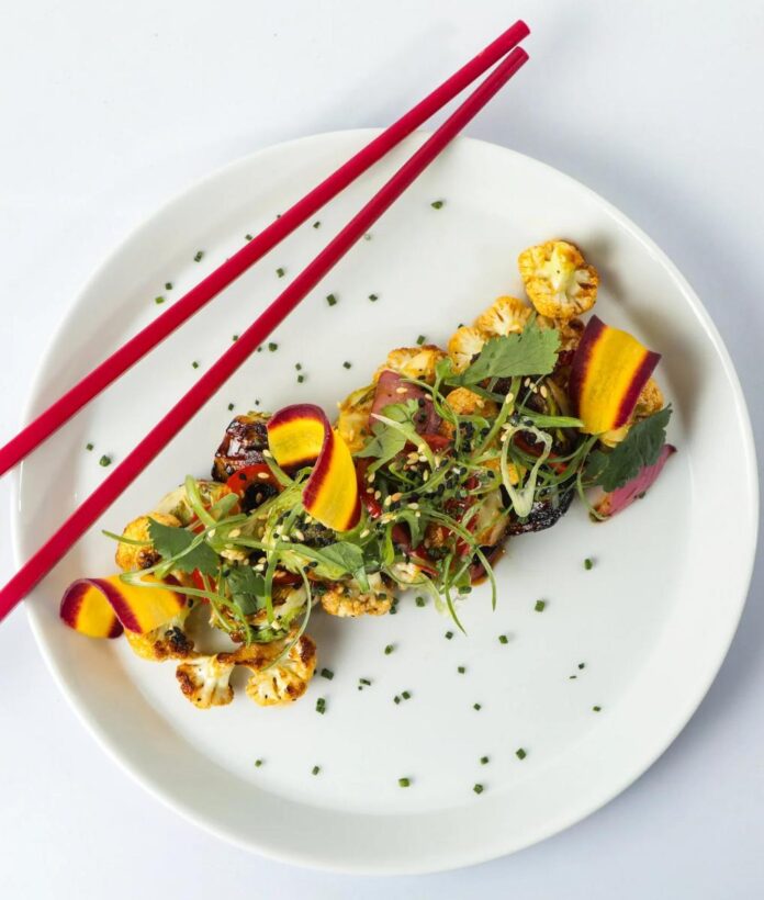 A plate of cauliflower stir-fry with sliced ginger, carrots, and green onion, with a pair of red chopsticks resting on the side.
