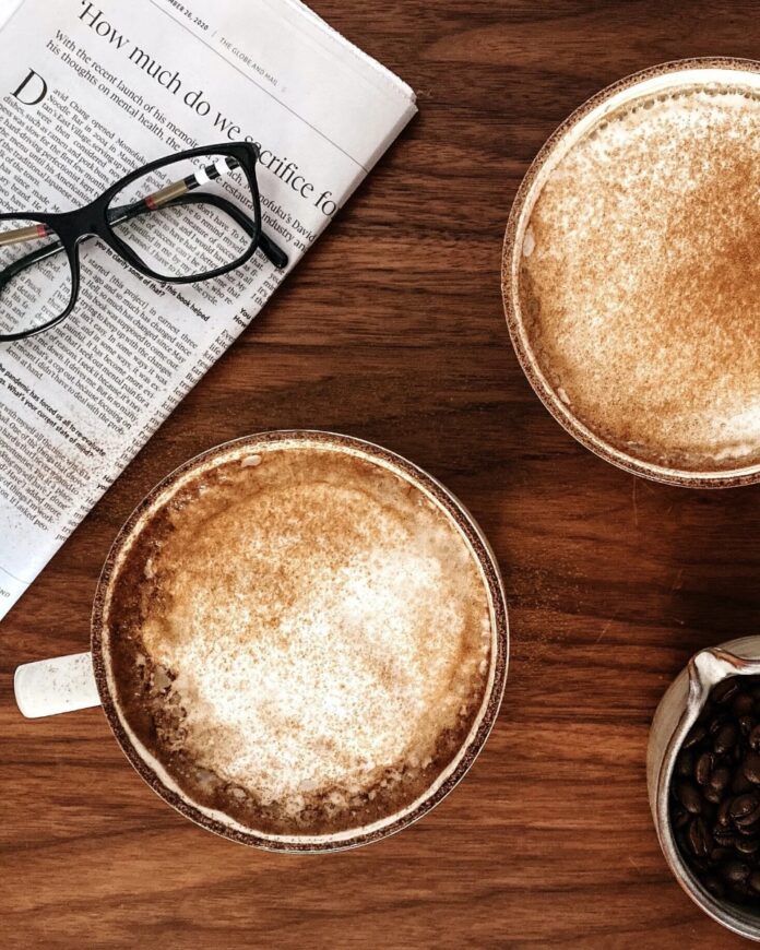 Two mugs of cinnamon spice lattes on a wooden table next to a newspaper and a small bowl of coffee beans.