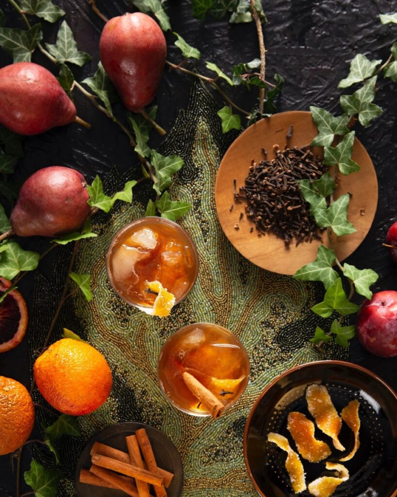 A Clove Old Fashioned cocktail in a rocks glass, garnished with a candied orange peel and a cinnamon stick, with cloves, cinnamon, and oranges on a beaded placemat.