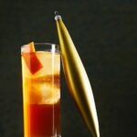 A rocks glass of drunk uncle cocktail with a large ice cube and an orange peel, next to a gold Christmas ornament.