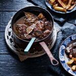 A large, copper pot of beef stew with bay leaves, a small plate of the stew next to a larger plate of french fries.