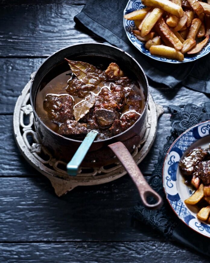 A large, copper pot of beef stew with bay leaves, a small plate of the stew next to a larger plate of french fries.