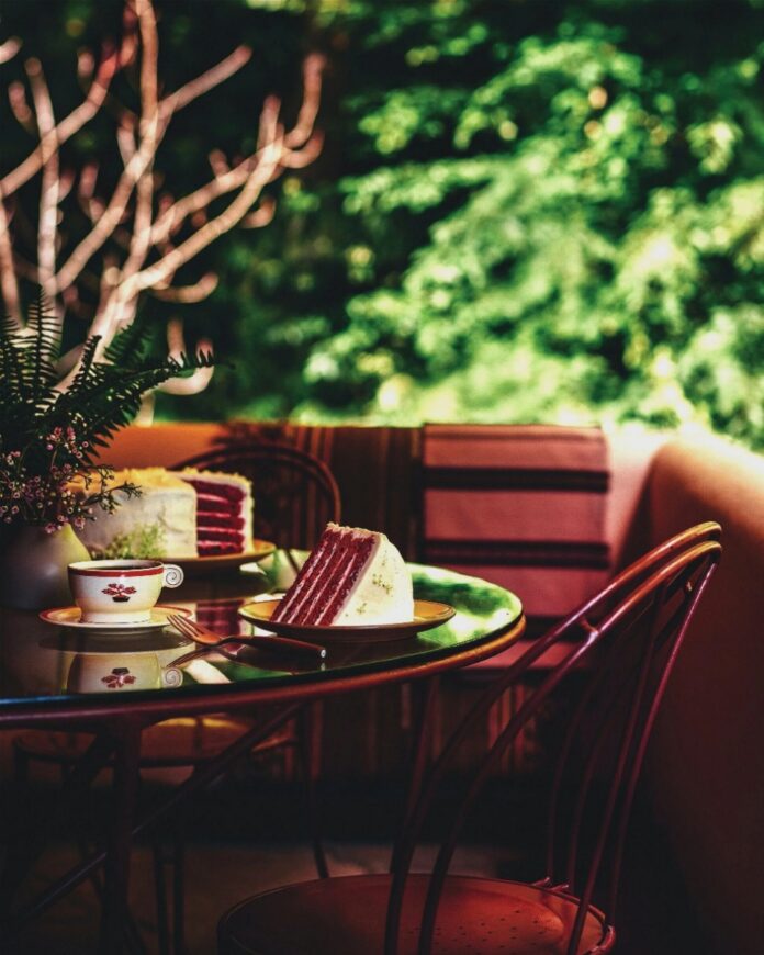 Edgar Sr.'s New Red Devil's Food Cake A glass-topped outdoor table with two slices of red devil's food cake on green plates, with a cup and saucer, and a plant in a white pot, against a soft green background of bushes.