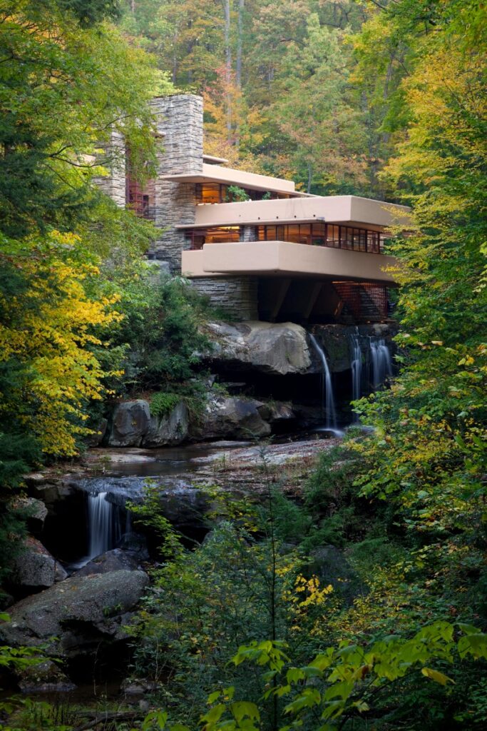 A long-shot, slightly elevated view of Frank Lloyd Wright's Fallingwater house, showcasing its tiered stone and stucco architecture over a waterfall, surrounded by lush, vibrant autumn trees.