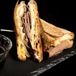 A halved French onion dip sandwich recipe made with leftover beef brisket, melted cheese and thinly sliced meat, resting on a black platter.