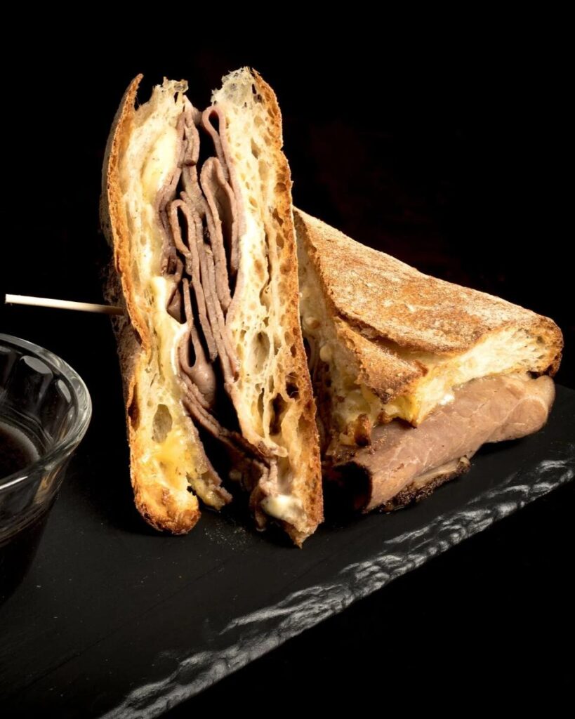 A halved French onion dip sandwich recipe made with leftover beef brisket, melted cheese and thinly sliced meat, resting on a black platter.