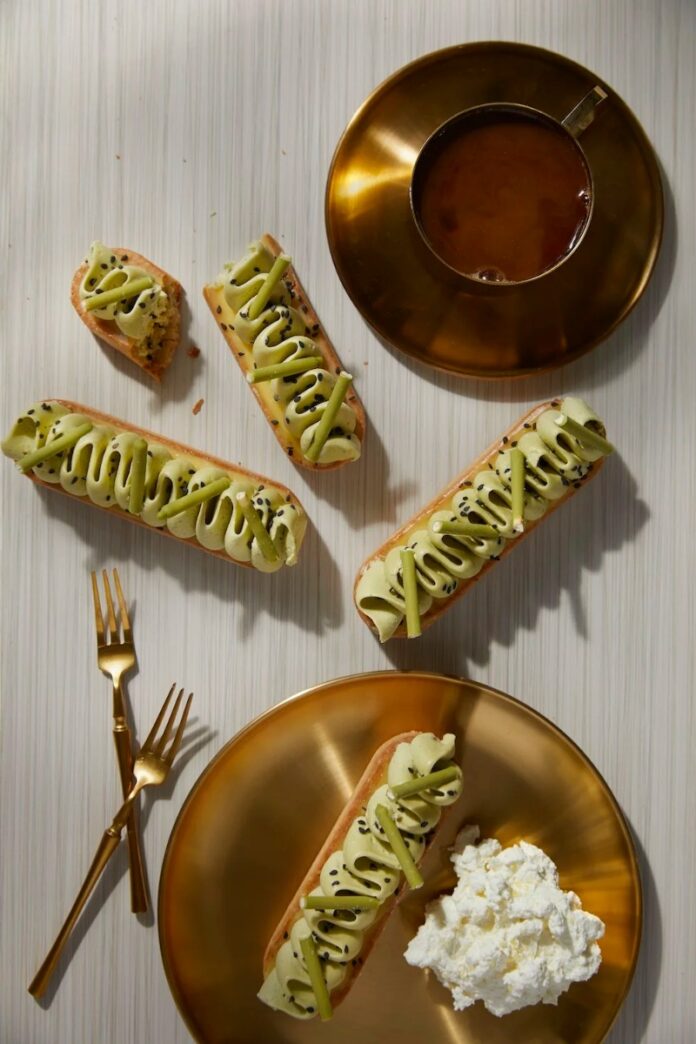 Four long tarts with green filling on a golden plate, with a coffee cup and two forks next to them on a white striped surface.