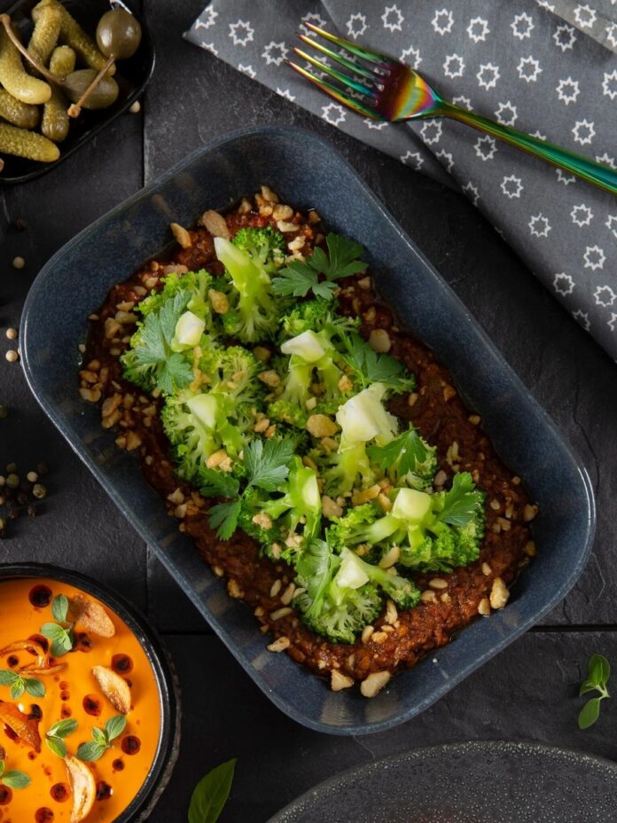 A plate of grilled broccoli with romesco sauce, topped with parsley and chopped nuts, with a small bowl of capers.