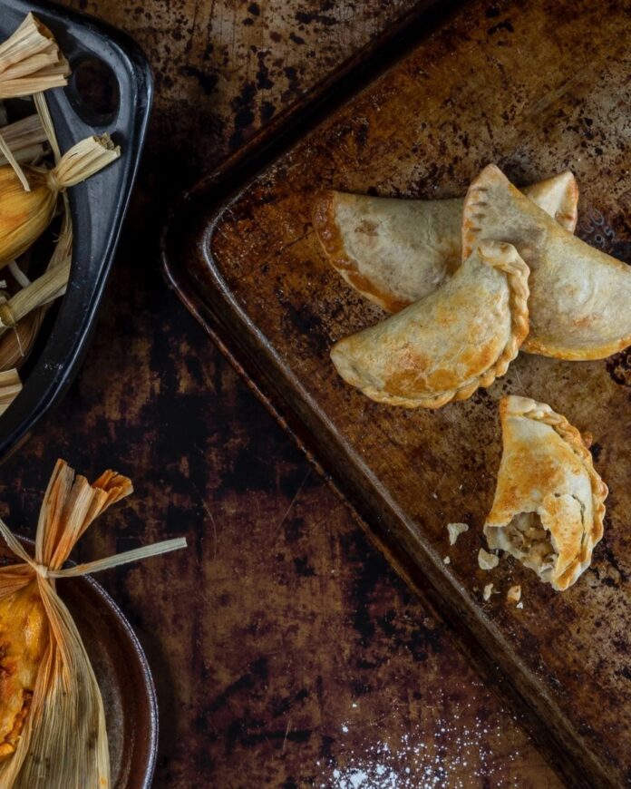 Several golden brown empanadas on a rustic metal baking sheet, with one broken open to show the filling.