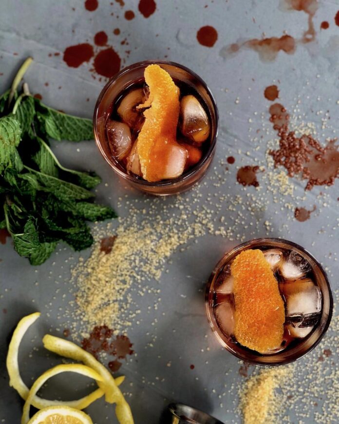 Two cocktails in rocks glasses garnished with large orange peels, next to sprigs of fresh mint and a messy pile of sugar and liquid splatters.