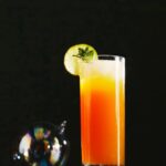 A tall glass of an orange and yellow kid's choice cocktail, garnished with a lime slice and mint, sits on a dark surface.