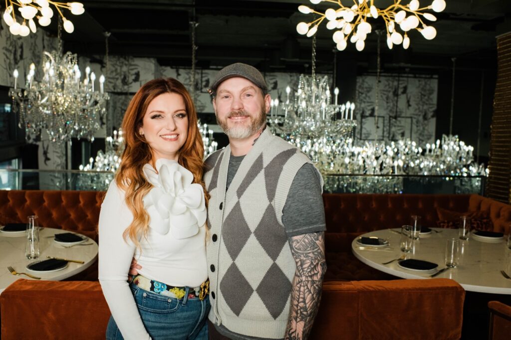 A man in a plaid sweater vest stands with a woman in white. 