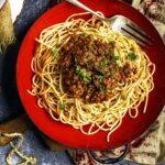 A red bowl filled with spaghetti topped with lentil bolognese sauce and parsley, with a fork resting on the side.