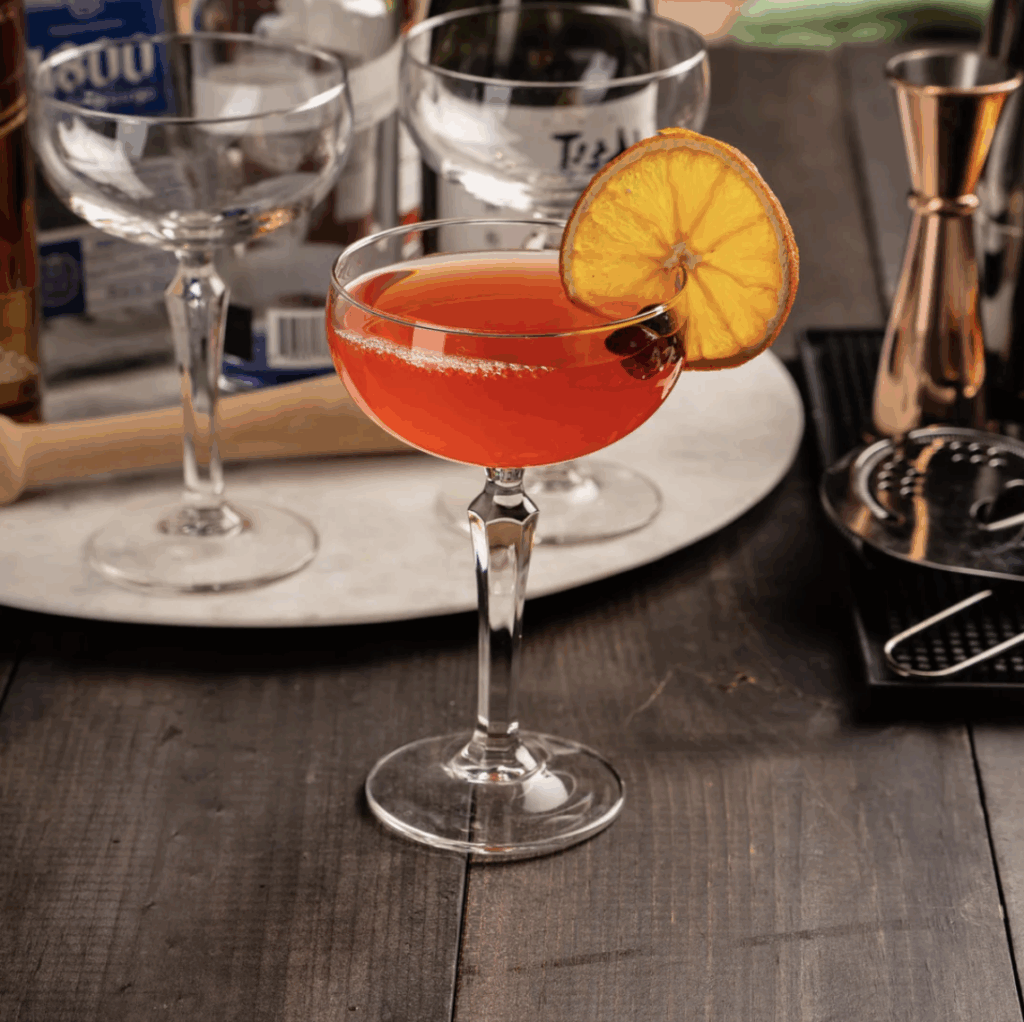 A coupe glass on a wood table with a red drink and orange wheel on the rim. 