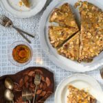 A glazed almond cake topped with sliced almonds and orange zest, with two slices on separate plates and a glass of amber liquid, all on a patterned white tablecloth.