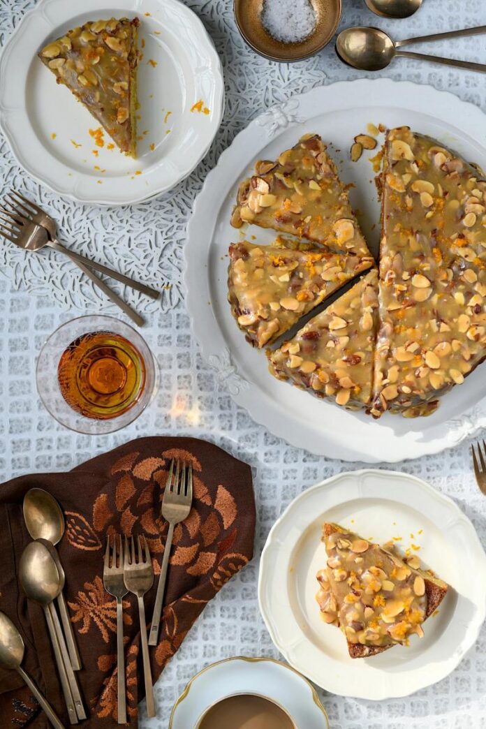A glazed almond cake topped with sliced almonds and orange zest, with two slices on separate plates and a glass of amber liquid, all on a patterned white tablecloth.