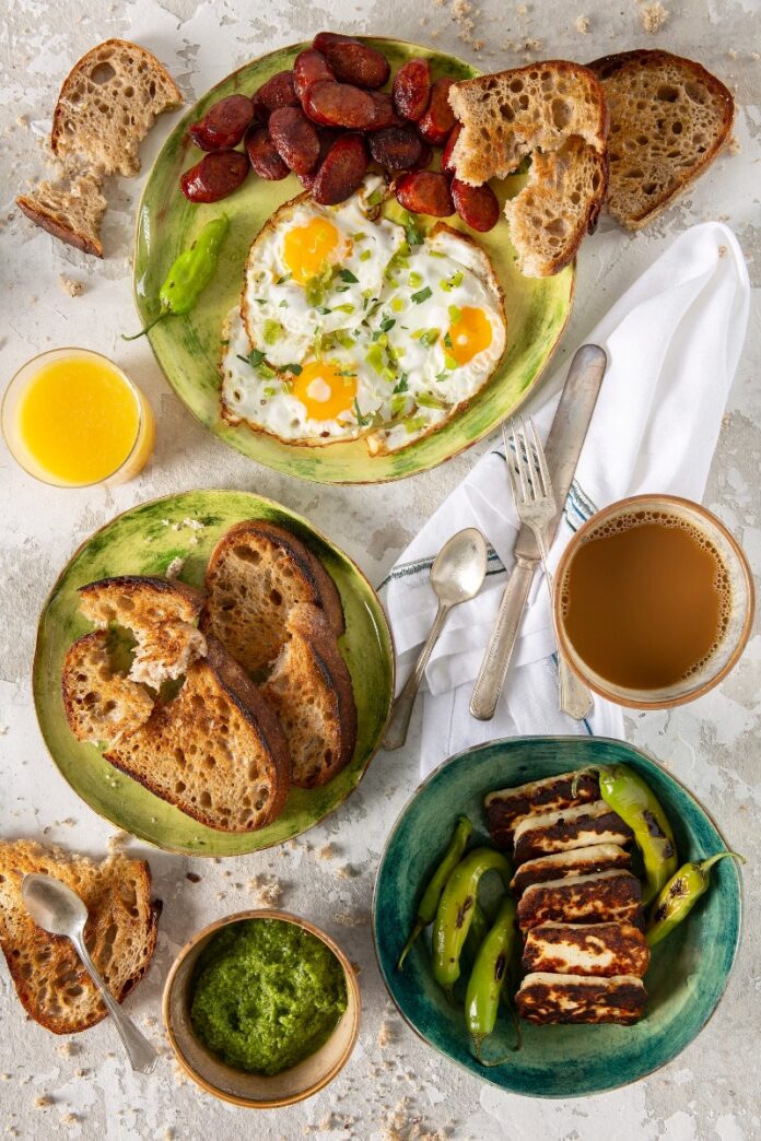 Mediterra's Fried Eggs A Spanish breakfast with fried eggs and chorizo, grilled halloumi cheese with green peppers, and bowls of zhoug sauce and orange juice, all served with toasted bread.