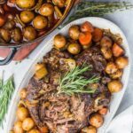 A slow-cooked beef brisket on a white platter surrounded by roasted potatoes and carrots.