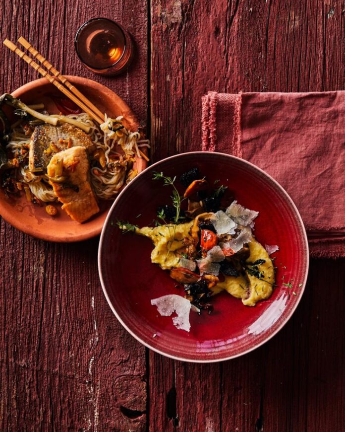 A plate of pan-seared trout with noodles and vegetables next to a red bowl of creamy polenta with wild mushrooms and shaved parmesan, all on a weathered red wooden surface with a red napkin.