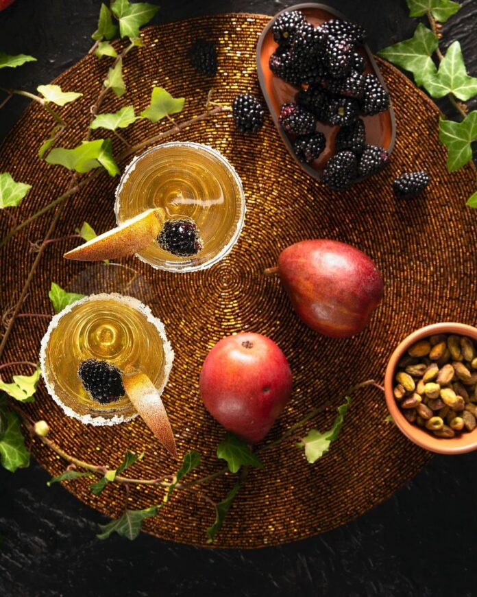 A pair of pear cider and gin bellinis in sugar-rimmed glasses, garnished with blackberries and pear slices, on a textured placemat.
