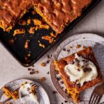 A baking pan of pumpkin blondies with a square slice topped with cream cheese frosting and chocolate shavings on a small plate.
