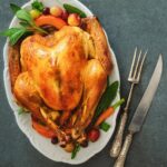 A perfectly roasted whole chicken on a white platter garnished with grapes, apples, cranberries, and fresh sage, alongside a vintage carving set.