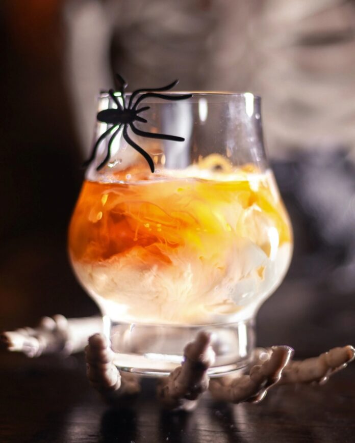 Sip and Scare A clear glass of a creamy, orange-hued cocktail, garnished with a plastic spider, held aloft by a skeleton hand against a dark, foggy background.