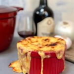 A single serving of French onion soup in a red crock with melted cheese overflowing the sides, a bottle of red wine and a wine glass, and a large onion and garlic clove in the background.