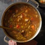 A large silver stockpot filled with a steaming broth of chicken, carrots, and celery, with a ladle resting inside.
