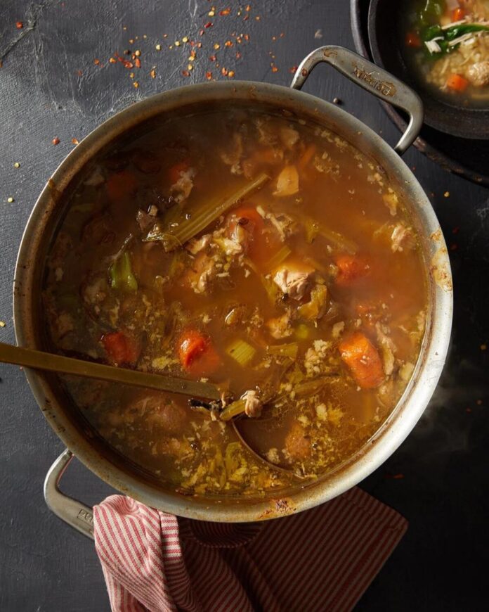 A large silver stockpot filled with a steaming broth of chicken, carrots, and celery, with a ladle resting inside.