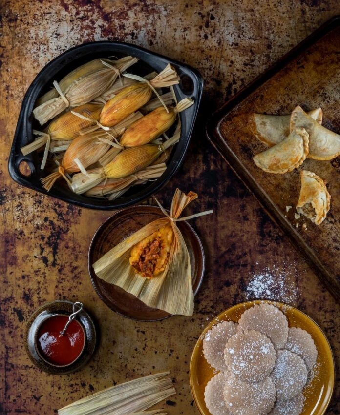 A black bowl filled with traditional tamales wrapped in corn husks, with one unwrapped tamale on a smaller plate showing a rich, red filling. Beside the plates are other New Mexican foods, including empanadas and biscochitos.