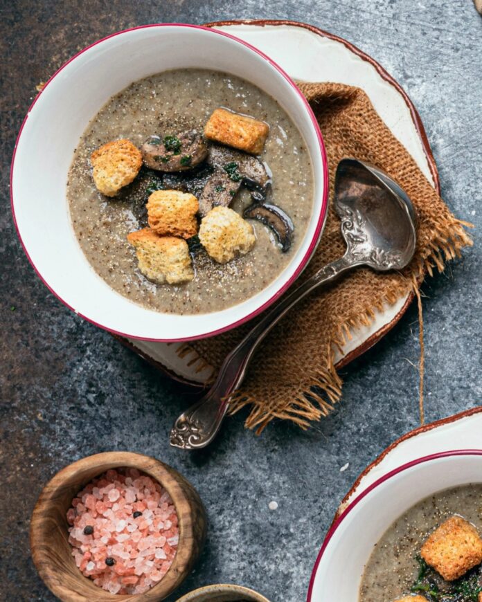 Vegan Mushroom Potato Soup A bowl of vegan mushroom potato soup garnished with croutons and sliced mushrooms, with a ladle resting on a brown napkin.
