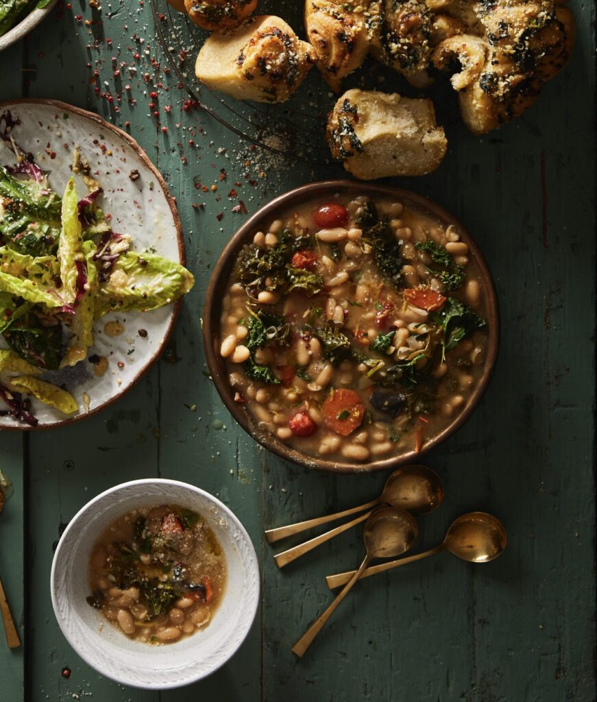 A green picnic table with a Caesar salad, garlic knots, and beans and greens bowl on top.