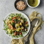 A panzanella mango salad sits on a white plate by bowls of seasonings and a set of silverware.