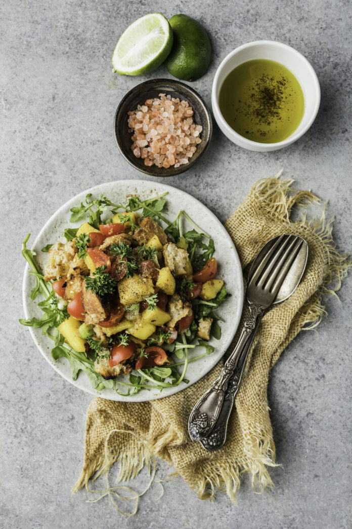 A panzanella mango salad sits on a white plate by bowls of seasonings and a set of silverware.