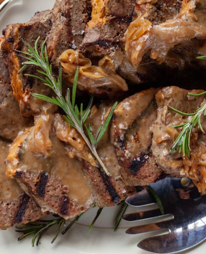 A close-up of grilled, sliced meatloaf topped with caramelized onions and brown gravy, garnished with fresh rosemary sprigs.