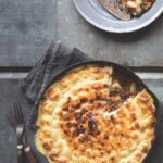 A cast iron skillet filled with Shepherd's pie topped with a golden-brown, bubbly rutabaga and potato mash, with a serving on a small gray plate.