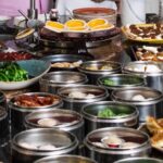 Various silver trays of dumplings sit in front of a tray of egg tarts.